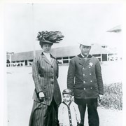 Three people are standing in front of stable area of Elias J."Lucky" Baldwin's Santa Anita Race Track.  L-R: Mrs. John (Ella) Ott; eldest son, Donald; and City Marshall, John Ott. (Their daughter told us in 1980 that everyone called her father Jack.) Mrs. Ott is wearing a lavishly feathered hat and a tailored suit.  Marshall Ott is wearing a dark double-breasted suit with his badge.