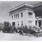 Arcadia City Council members in a decorated Hupmobile Touring Car parked on Huntington Drive next to City Hall.  Behind is a fire truck, also decorated, with 10 firemen on the truck or near by.  The man at the wheel of the fire truck is Jim Nellis.  The Councilmen are: Rear seats,L-R: Ferd E. Gram; Arthur N. Multer; and Charles Hawk. Front seat,L-R: Samuel L. Wheeler; John T. Joyce, the Hupmobile Dealer.  Fifth member of Council, John Granville was not present.  Seated at the base of the pillar of City Hall is Adrian Winkler and standing beside him is George Newton.  The vehicles are on their way to dedication of new concrete span over Santa Anita Wash.