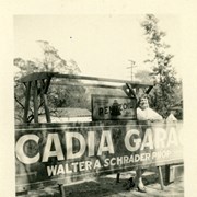 Woman (probably Nell Schrader) standing behind large sign reading ARCADIA GARAGE, WALTER A. SCHRADER PROPRIETOR.  There is also a sign advertising Penzoil for sale.