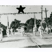 Rancho Santa Anita float entry for Rose Parade.  Two men in front wearing white, carrying poles with garlands and a red star in the middle. Float with red star visible in background. This entry was designed by Anita Baldwin to reflect her involvement with the Red Star Society.