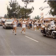 Two Olympic torchbearers on Santa Anita Avenue as part of the Olympic Torch Relay for the XXIII Olympiad in Los Angeles. Motorcycle police escort also visible.
