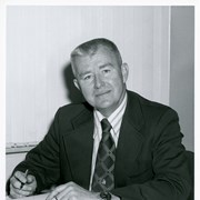Photo of Arcadia Police Chief Charles Mitchell, shown seated at a desk or table, wearing a suit and tie.  He has a pen in his right hand and has an open book in front of him.