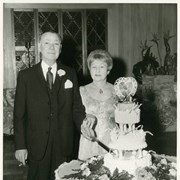Photo of Charles and Billie Eaton at what might have been a 50th wedding anniversary celebration.  The Eatons are dressed in formal attire, standing behind a 3-tier cake.  The Eatons are holding a cake knife and looking toward the camera.