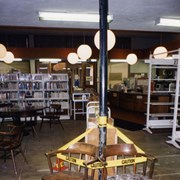Photo taken of Arcadia Public Library, 20 W. Duarte Road, during the 1995/96 expansion/remodel project.  This view is of the temporary adult reading area, looking toward the temporary circulation desk and temporary front entrance. In the center of the photo, a drainage pipe is surrounded by chairs and booktrucks.  This pipe was installed to help drain rain from the roof.  The carpet in this area was removed because of extensive water damage. Globe lights.