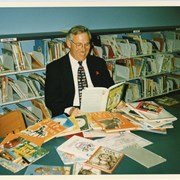 Councilmember Gary Kovacic is seen seated at a table in the Jerry Broadwell Childrens Room.  He is surrounded by books on the table and is looking at one of them.  Photo was taken by Dorothy Denne of the Arcadia Weekly to be used in a display for National Library Week.