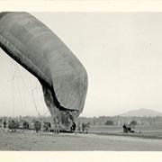 View west. Balloon being held for a lowering or launching maneuver. A small section of white fence shows on left. About 24 men in photo.