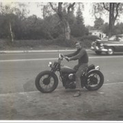 Man seated on motorcycle with cigarette in his mouth.  He is looking toward camera.Handwritten note on back of photo reads: "SW corner Duarte Rd & Golden West.  Robert D. Brannen on motorcycle.  Greer Residence in background left - Meyers house background right."