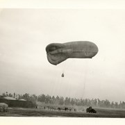 Photo of balloon apparently descending with men in basket suspended from beneath it.  Group of buildings and 2 tents show on left side of photo. Approximately 16 men involved in getting balloon down.  Truck also in photo.