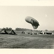 Photo shows 2 tents on left side of photo.  Balloon approximately 25 feet from ground.  Group of men involved in maneuver also seen.