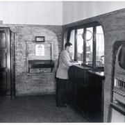 Man standing at dispatcher window in the Arcadia Police station at First Avenue and Wheeler Street. Door with telephone sign above it, First Aid sign on wall and cigarette machine near counter.