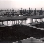 View of front door of one-story Arcadia Police Department building at 250 W. Huntington Drive. The building has a flat roof. Man in suit stands inside the front door. Parking lot of the Santa Anita Racetrack in background.