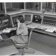 Interior Arcadia Police Department building at 250 W. Huntington Drive. Female employee in APD uniform, possibly a dispatcher, sitting at a telephone switchboard and communications panel, using rotary dial telephone and writing. Also seen are a manual typewriter, microphone, and the time