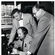 Interior Arcadia Police Department building at 250 W. Huntington Drive. Three men at the same dispatch or switchboard communication station as shown in photo #1753. One man in APD uniform, sitting with headset on, two men in suits look over him.