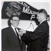 One man pins an Arcadia police badge on another man's left lapel. A pennant on the wall reads Arcadia Mounted Police.