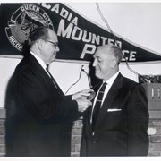 Man wearing glassespins an Arcadia police badge on another man's right lapel. A pennant on the wall reads Arcadia Mounted Police.