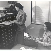 Female employees of the Arcadia Police Department, wearing badges. Standing is Flora Mae Keeville, the Chief's secretary September 1956-March 1959, flipping through a file cabinet. Sitting is Mary Desmond, a steno clerk from October 1955-November 1958, at a manual typewriter.