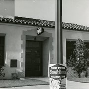 Entrance to Arcadia police station at 50 Wheeler St. Neon "Police" sign above doorway. Red clay tiled roof. Sign posted on pole reads "Thrift Twins Save-A-Rama Celebration July 1-6. Presented by Downtown Arcadia Merchants."