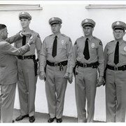 Four unidentified male police officers lined up, along an exterior wall, in uniforms and hats. Man in suit is pinning a badge on the first officer on the left.