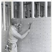 Construction in progress of the one-story Arcadia Police Department building at 250 W. Huntington Drive. Man with hammer nails piece of gypsum to a stud in the frame. Man holds nails in his mouth. Text on the gypsum reads "Kaiser Gypsum."