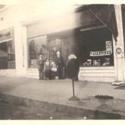 Seaquist's Drug Store.  Pictured in front are three people: younger man in light suit, woman seated, and older man in dark suit.  Among other signs in the window is one for polo at Midwick Country Club which was between Alhambra and Monterey Park.  On side of building is painted ad for Wrigley's P.K. chewing gum.  Located about 200 block of N. First Avenue.