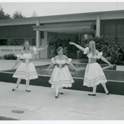 Three girls wearing identical dress costumes and ballet shoes, in dance poses outside Arcadia City Hall. There is a fountain behind the girls that no longer exists. The City Hall tile mosaic wall (mural) depicting Arcadia history, by artist Jay Rivkin, is seen in the background (noted November 27, 2013-the murals had been covered by stucco for many years but were restored this year).
