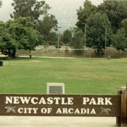 Sign reads "Newcastle Park City of Arcadia." View of  trees, volleyball nets, and picnic tables.