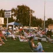 Arcadia Recreation Department summer concert at First Avenue School front lawn, shows a view of the audience seated on lawn chairs. The street in the background is First Avenue. A "Soft Water Service" business is seen in the background. Although the back of this photograph is stamped with its processing date of August 1997, "Summer Concert 1996" was handwritten on the envelope it came in from Arcadia Recreation Department.
