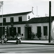 Early 1950s exterior view of Arcadia police and fire department building at Wheeler Avenue and First Avenue. Two police officers are sitting on their motorcycles, parked by the curb. The doorway has a "Police" sign above it.