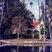 Queen Anne Cottage at the Los Angeles County Arboretum, with its distinctive red roof and bell tower. View of the house is partially obstructed by trees. The lake is in the foreground.