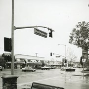 Downtown Arcadia intersection of Huntington Drive and First Avenue showing the building on the northeast corner, photo taken from the southwest corner (the corner of Washington Mutual bank). A Chevron gas station on the southeast corner, hidden by trees, showing gasoline prices, and cars on the street. Also, bench and traffic lights. Photo by Terry Miller.