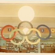Children's Room, Arcadia Public Library, 20 W. Duarte Rd. decorated for Summer Reading Club with 1968 Summer Olympics theme, featuring Olympic rings, flags and emblems of various nations. Shows interior furnishings and details such as card catalog, globe pendant lights, file cabinet, seating, shelving, staff person at a rounded information desk.