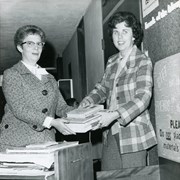 Volunteers handling materials in the library behind a book truck. Jean is wearing horn-rimmed glasses and Doris is wearing a plaid jacket. Left-Right: Jean Livingston, Doris Selmer. Friends of the Library.