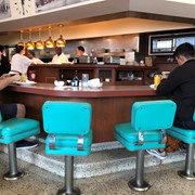 Interior view of Rod's Grill showing customers eating at the counter, sitting in colorful turquoise seats. Waitresses and cooks can be seen in the kitchen area, as well as a portion of two of the historic photos that the restaurant displayed on its walls. Rod's was located at 41 W. Huntington Drive in Arcadia, along Historic Route 66, from 1957 through February 2023.
