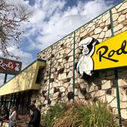 Exterior view of Rod's Grill showing the south side of restaurant with its rock mosaic wall and both the sign on the front of the building and the pole sign bearing its name  that stands on the west side. Customers can be seen waiting outside. Rod's was located at 41 W. Huntington Drive in Arcadia, along Historic Route 66, from 1957 through February 2023.