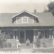 Neat clapboard house, taken looking straight on.  There are broad front stairs and solid wall enclosing porch.  There is a woman and a man sitting on front stairs.  House was located at NW corner of Colorado and First Avenue.
