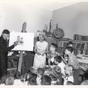 Author and illustrator Leo Politi talks to children in Children's Room, Arcadia Public Library, 25 N. First Avenue.  Children's Librarian Marjorie Hickerson (later, Phelps) is standing in white dress by easel.