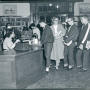 Group of high school students lined up to check books out in the evening. Grace Rahm is clerk seated at desk helping them.  In background are 3 additional patrons browsing.