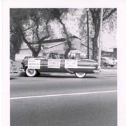 Vi Chaffers Mahr at the wheel of a convertible decorated for Arcadia Public Library's entry in Arcadia's 50th birthday party parade. San Gabriel Valley Lumber Company building is visible in the background. Location is approximately First Avenue and Wheeler Street.