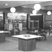 View west across children's room toward book stacks.  Regular tables and chairs are in place, and record listening tables. Globe lights.