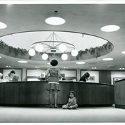 View of circulation desk under skylight dome of Arcadia Public Library. In view, working at desk, are Freda Bernard on left with glasses and Carole Schubert (later, Wilson) on right.  Two women patrons are being helped by Freda Bernard, and one woman with small girl waits at center. Globe lights and "wagon wheel" fixture.