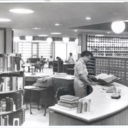View of reference desk (originally located in center of area just inside doors of adult reading room). Card catalog at this time ran north and south.  Both Reference Desk and catalogs were relocated in 1972. Alfreda Bolduan, head of Reference Department, is standing at desk. Rounded desk, globe lights, telephone, and typewriter visible.