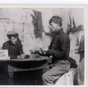 Two young men playing cards at a table in a room.  Man with cap is Richard Herman Shierske, who was a jockey for E.J."Lucky" Baldwin. He was licensed under the name of Richard Herman.  Other young man is Charles McIver.