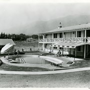 Portion of the exterior of the guest accommodations at the Westerner Motel. Photo shows swimming pool, with people lounging about.  The Y at intersection of Colorado Place and Colorado is visible as well as the San Gabriel Mountains.
