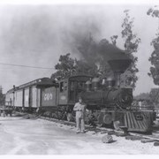 Train on track with William Parker Lyon standing alongside, outside Pony Express Museum.  Engine has Eureka, Nevada painted on it.  There are two cars attached to engine, one appears to be a baggage car, the other a passenger car.  Note: on left side of photo a portion of Santa Anita Racetrack Clubhouse is shown. Engine #7 of the Eureka Palisades Railroad, in use in 1875, was salvaged from Palisades, Nevada.