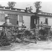 William Parker Lyon, owner of Pony Express Museum, shown standing between two old fire engines in outside exhibit at the museum.  Fire engines are standing in front of two cars of Narrow Gauge R.R.  Train has painted on it: Eureka, Nevada Narrow Gauge.  Mr. Lyon's shirt has insignia bearing letters L.E.