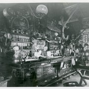 William Parker Lyon serving drinks behind bar located in one section of Pony Express Museum. (It did not operate for the public.)  Behind the bar are many different bottles and kegs.  There appear to be gaming tables in this room also.  Note: spittoons under bar railing.