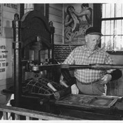 William Parker Lyon, owner of Pony Express Museum, dressed in plaid shirt with black sleeve protectors common to the trade of the printer. He is standing beside an old printing press.  Behind him can be seen the compartments which held the type.