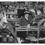 William Parker Lyon, owner of Pony Express Museum, pictured behind the counter of what seems to be a ladies apparel store.  He is wearing a ladies hat with a large feather on it.  On shelves behind him are many boxes designed to hold merchandise.