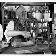 William Parker Lyon, owner of Pony Express Museum, dressed in paisley vest with white shirt. He is using a juice or drink mixer with a hand crank, with two glasses (two drinks).