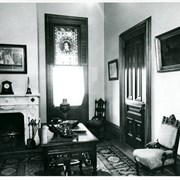 Interior view of Queen Anne Cottage.  View shows marble fireplace with sailing ship hanging over it.  There is a stained glass window with a head shown in a circle.  Painting of a horse shown on wall on the right.